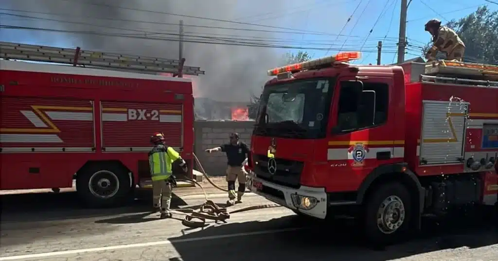 Incendio estructural en sector El Tejar de Quilimarí, con Bomberos trabajando en el control del fuego junto a carros y mangueras, mientras una vivienda arde al fondo.
